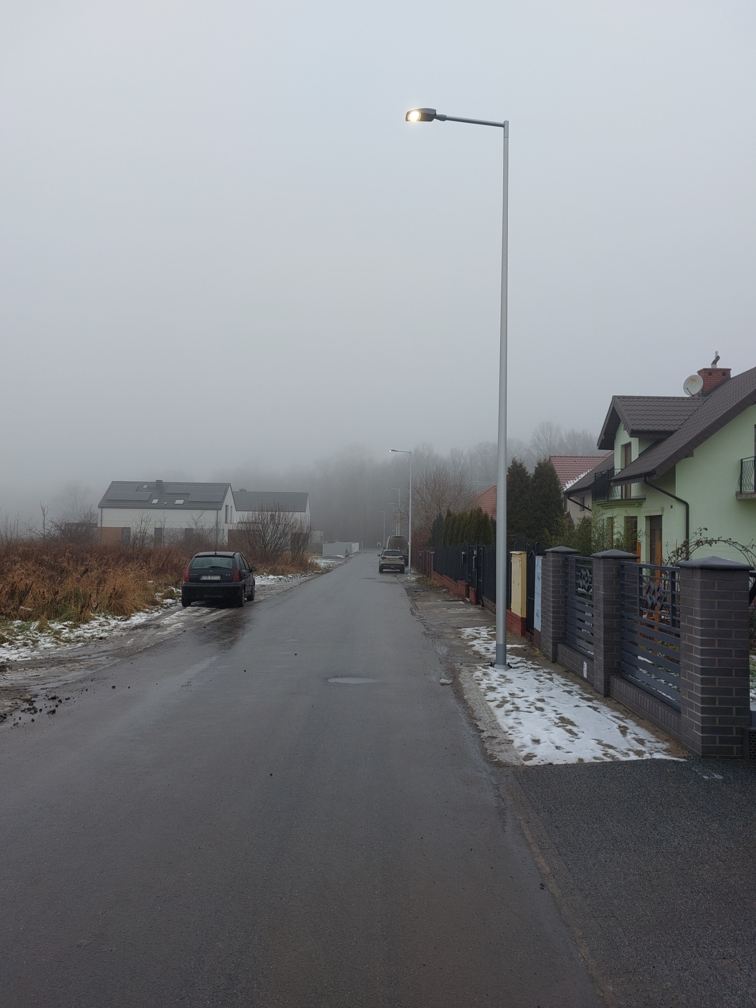 Mglisty krajobraz podmiejski z oświetloną uliczną lampą LED, mokrą jezdnią, częściowo ośnieżonymi poboczami i domami w tle.