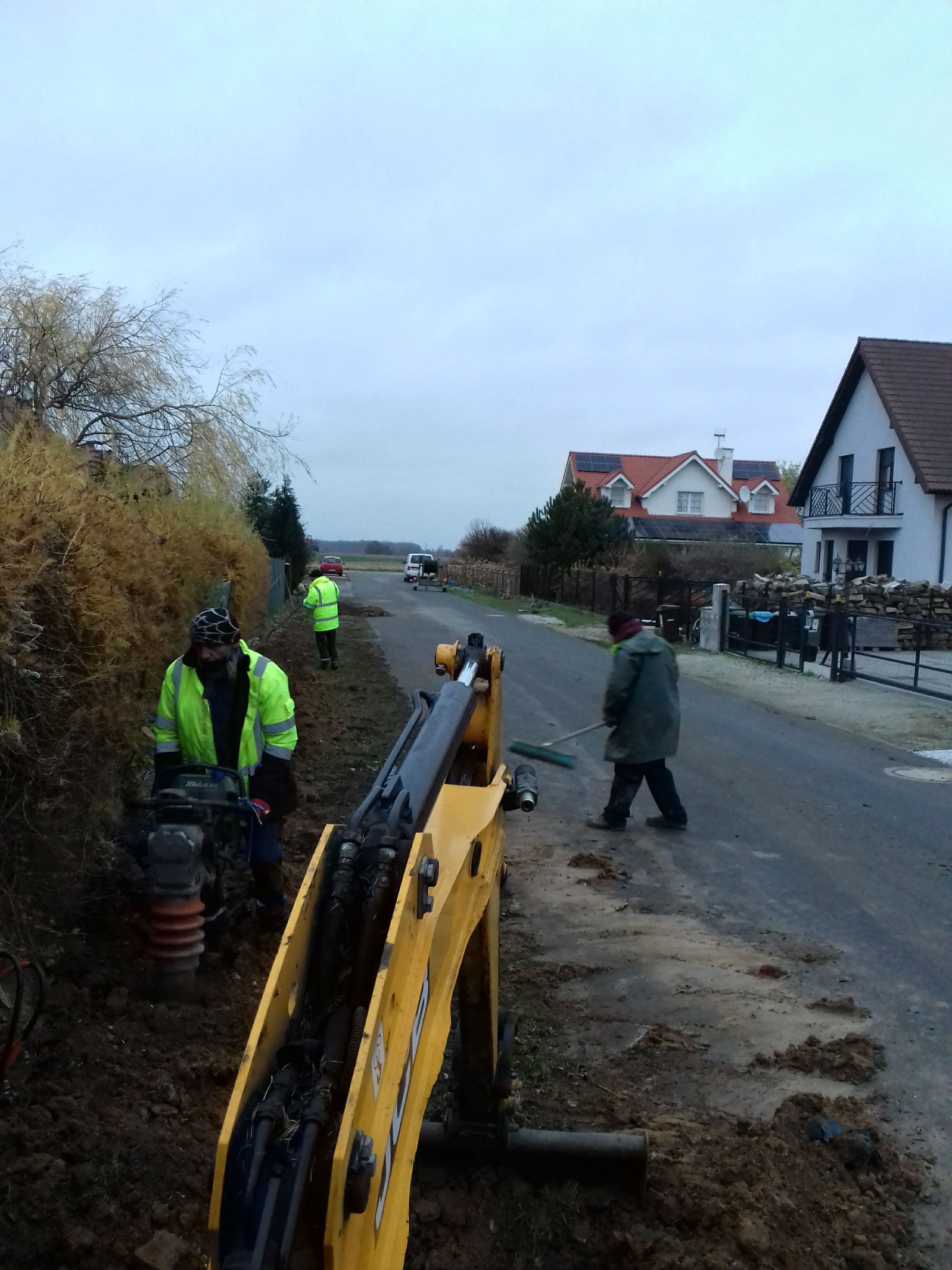 Pracownik zagęszcza ziemię ubijarką skoczną obok żółtej koparki, drugi sprząta pobocze miotłą, w tle zabudowa jednorodzinna i pochmurne niebo.