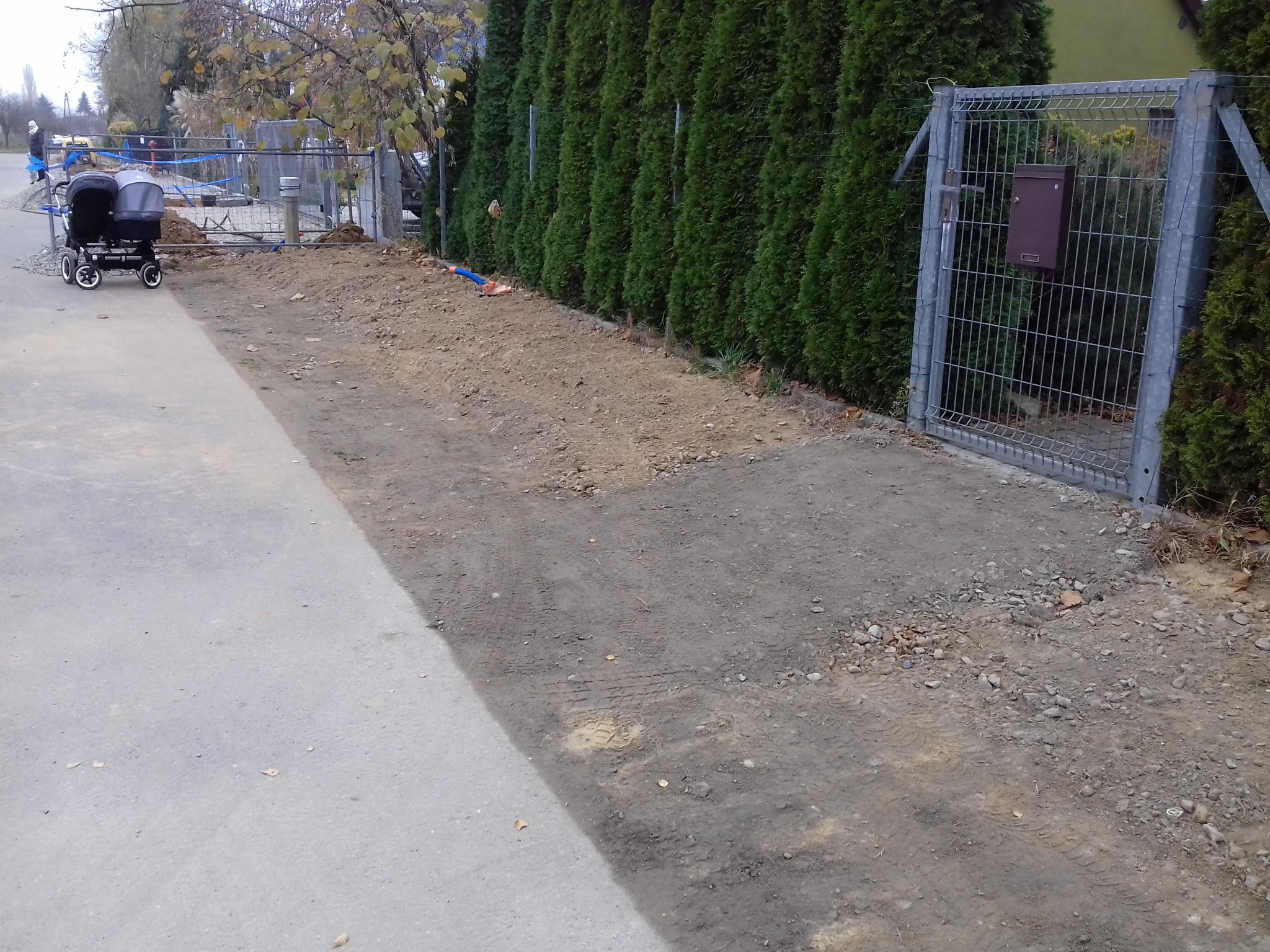 Przygotowany teren pod budowę z widocznymi śladami opon, ogrodzeniem z siatki i rzędem tuj w tle, w pobliżu zaparkowany wózek dziecięcy.