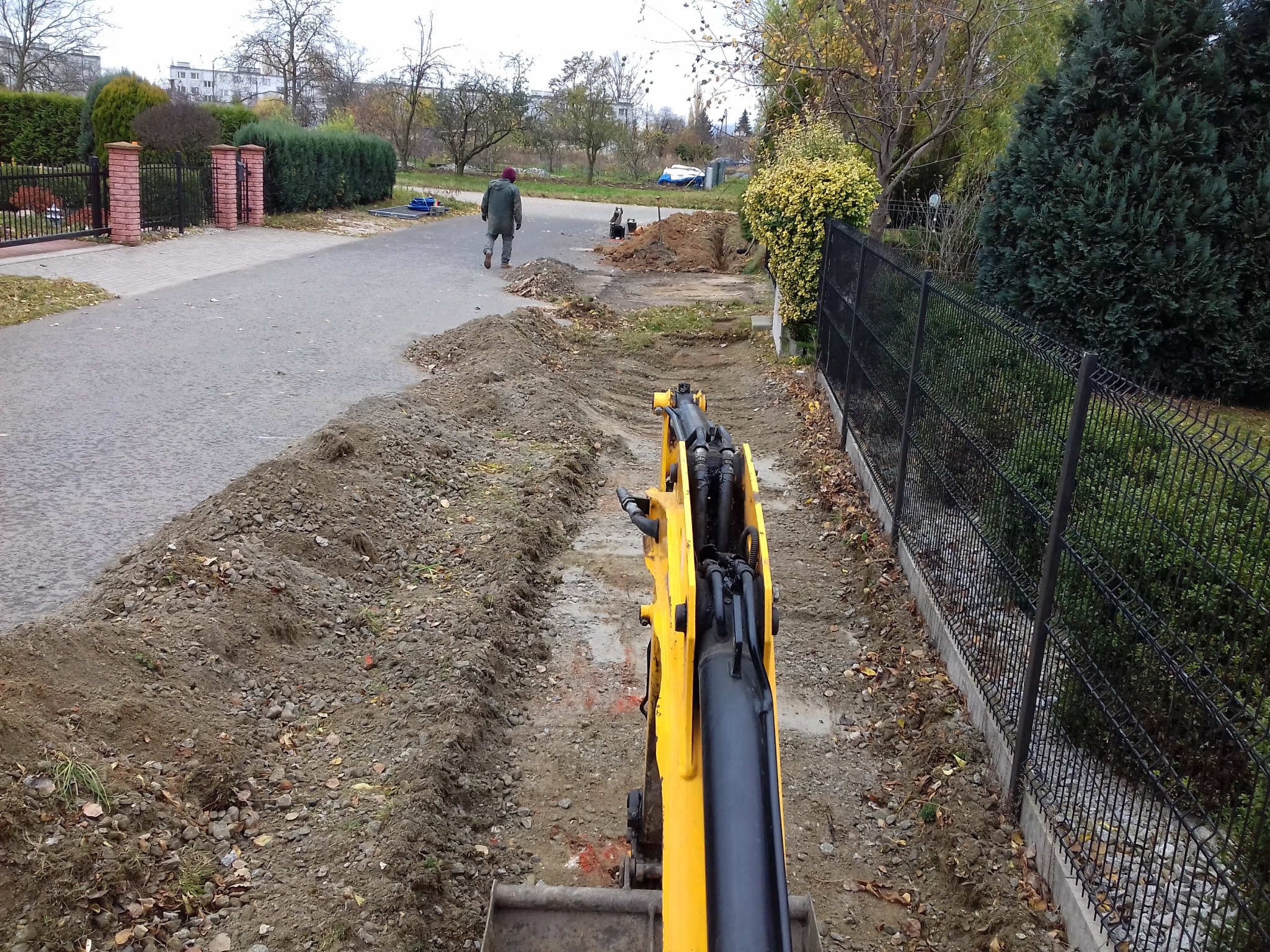 Żółta koparka na działce, wykopany wąski rów wzdłuż ogrodzenia z siatki, w tle ulica i spacerujący mężczyzna.