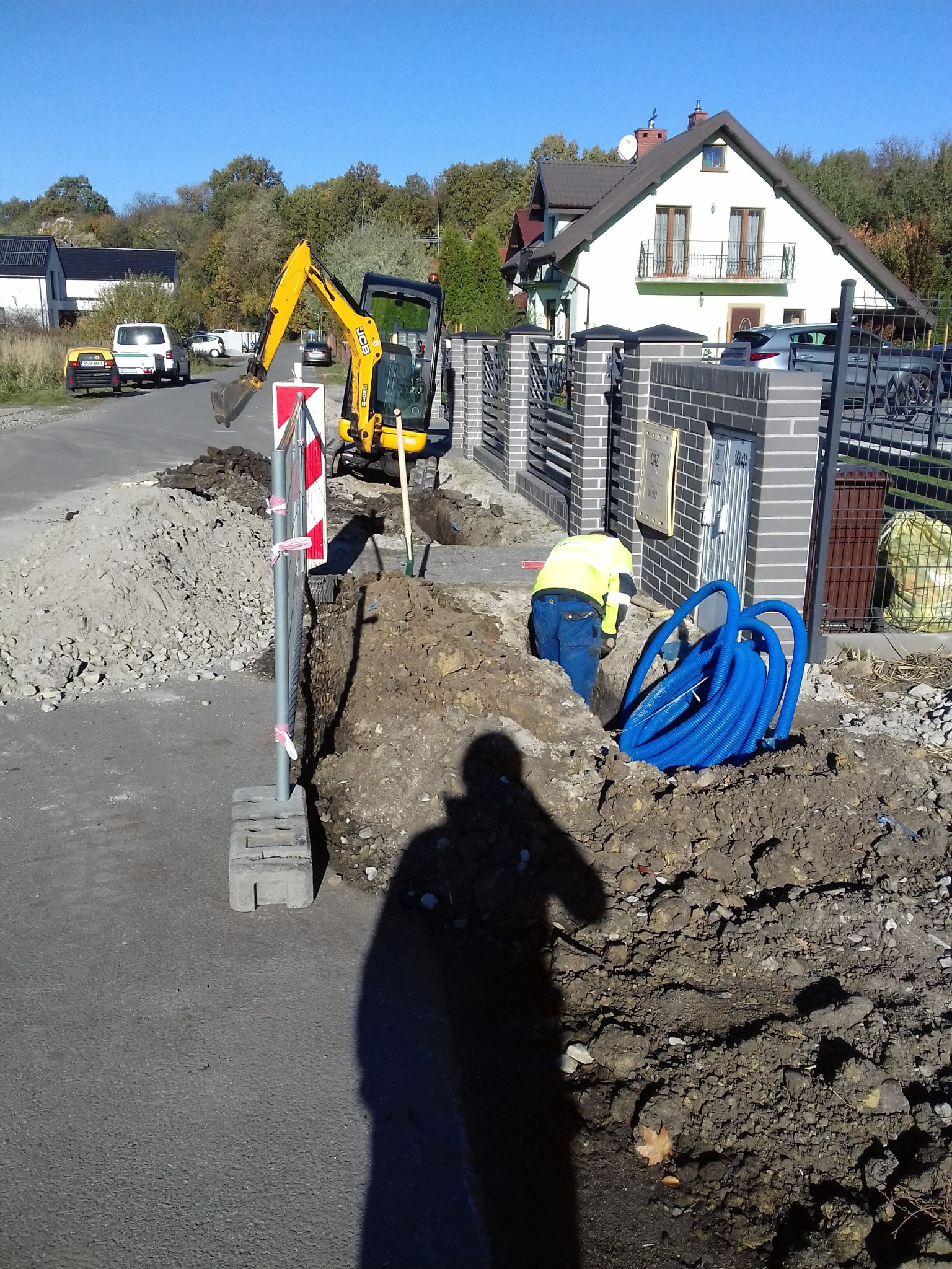 Pracownik w żółtej kamizelce układa niebieskie rury w wykopie obok ulicy, z mini koparką JCB w tle i stosem gruzu po lewej stronie, na tle domu jednorodzinnego.