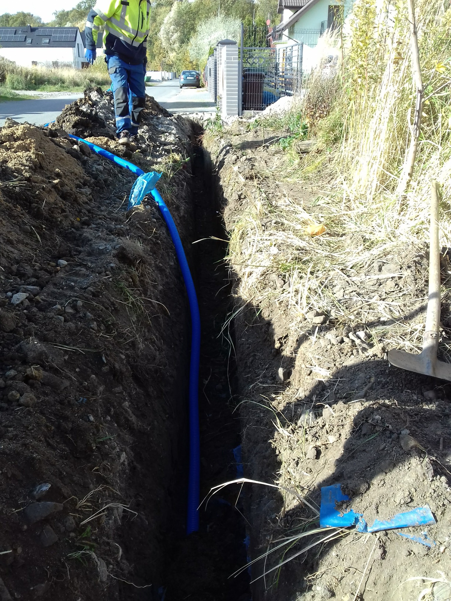Wkopana w ziemię niebieska rura karbowana, widoczna w wykopie, z pracownikiem w odblaskowej kurtce w tle, częściowo zasypana ziemią.