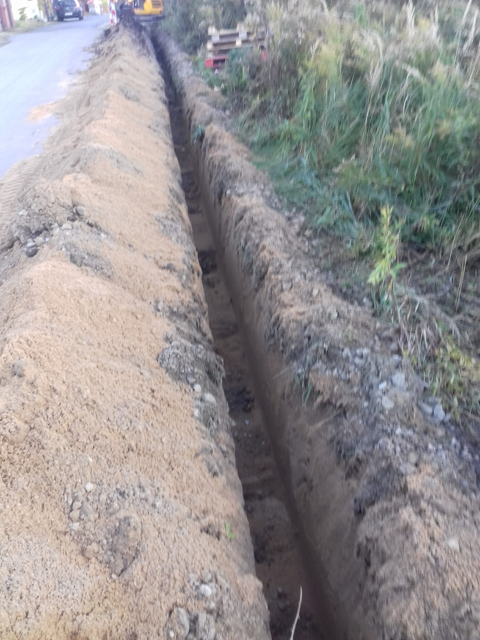 Długi, wąski wykop w ziemi, widoczny żółty sprzęt budowlany w tle, palety z materiałami budowlanymi na skraju terenu.