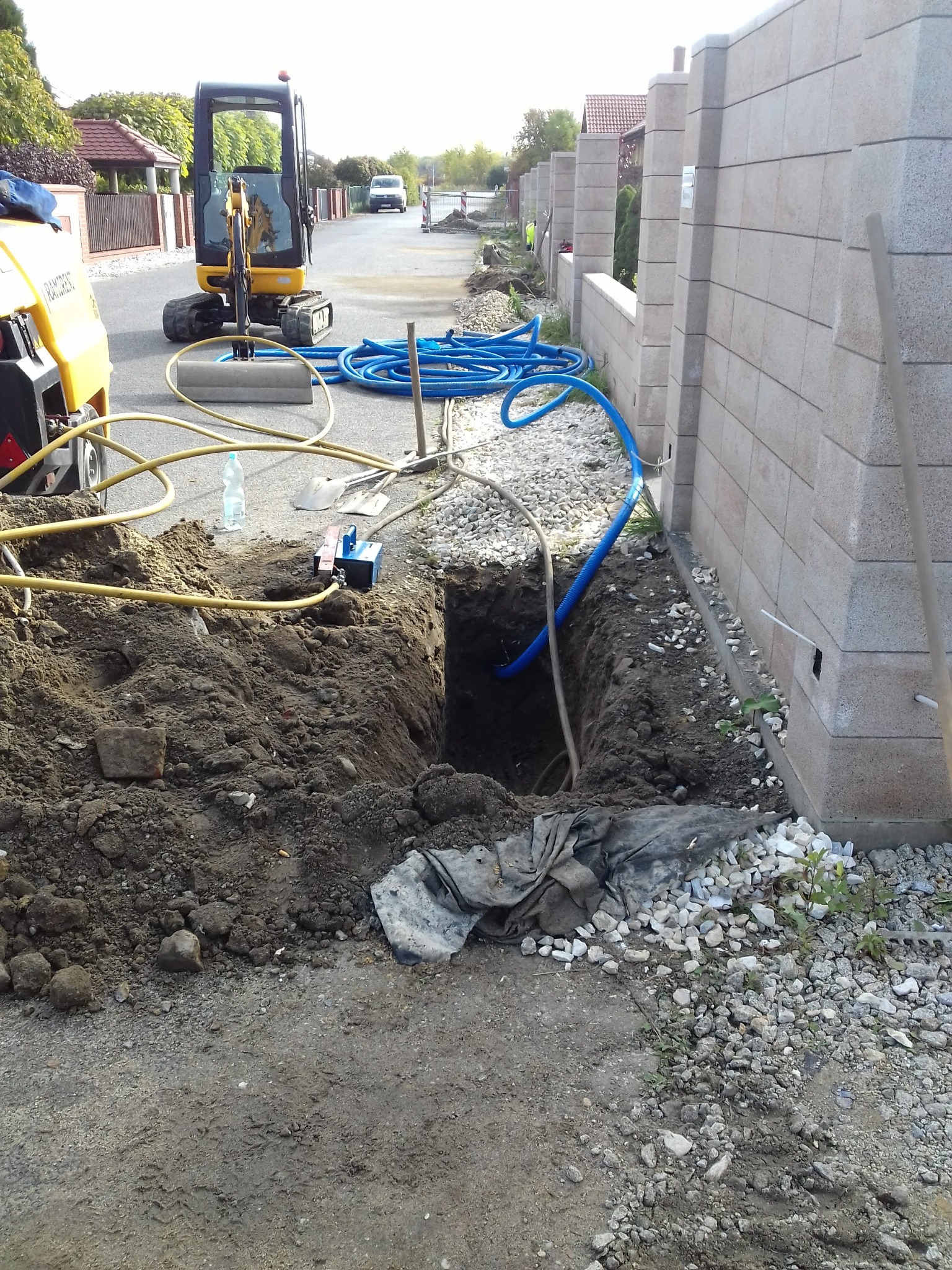 Wykop w ziemi z widocznymi niebieskimi rurami, żółtą rurą, butelką wody, łopatą i małą koparką na tle ulicy i betonowego ogrodzenia.