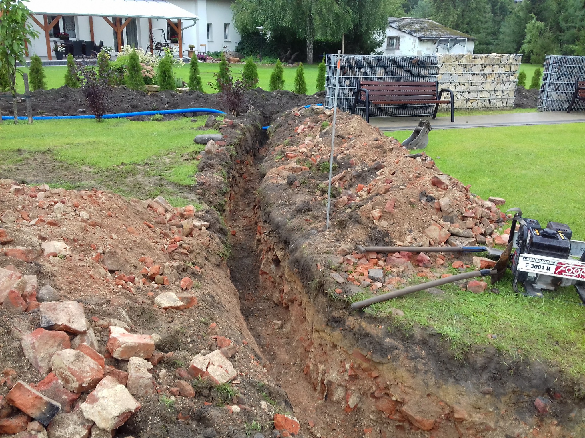 Wąski wykop w ziemi z widocznymi fragmentami cegieł, niebieska rura w tle, obok trawnik, w oddali dom i gabiony z ławkami.