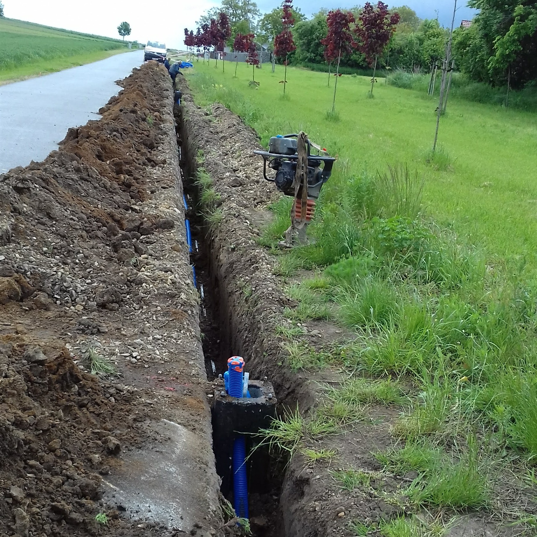 Wykop liniowy z ułożonymi niebieskimi rurami karbowanymi, widoczna studzienka rewizyjna z pomarańczowo-niebieską pokrywą, w tle ubijarka wibracyjna i pracownik przy wykopie.