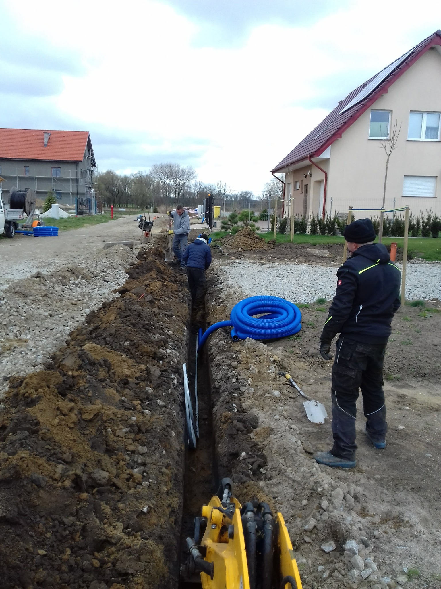 Wykop pod instalacje w trakcie prac ziemnych; widoczny wąski rów, ułożone rury instalacyjne, pracownicy, mini koparka i domy w tle.