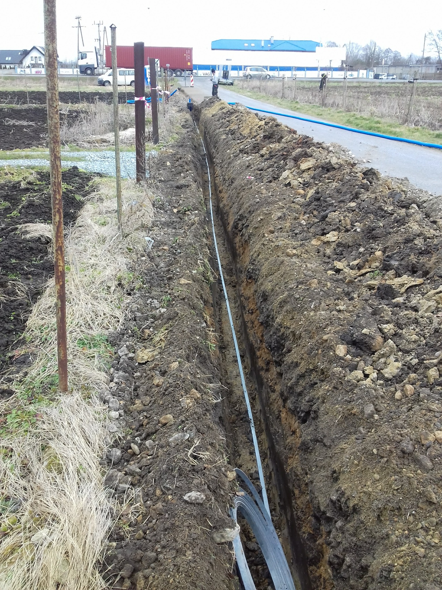 Wykopany wąski rów w ziemi z ułożonym szarym przewodem, widoczne ślady świeżo usypanej ziemi, w tle droga i zabudowania.