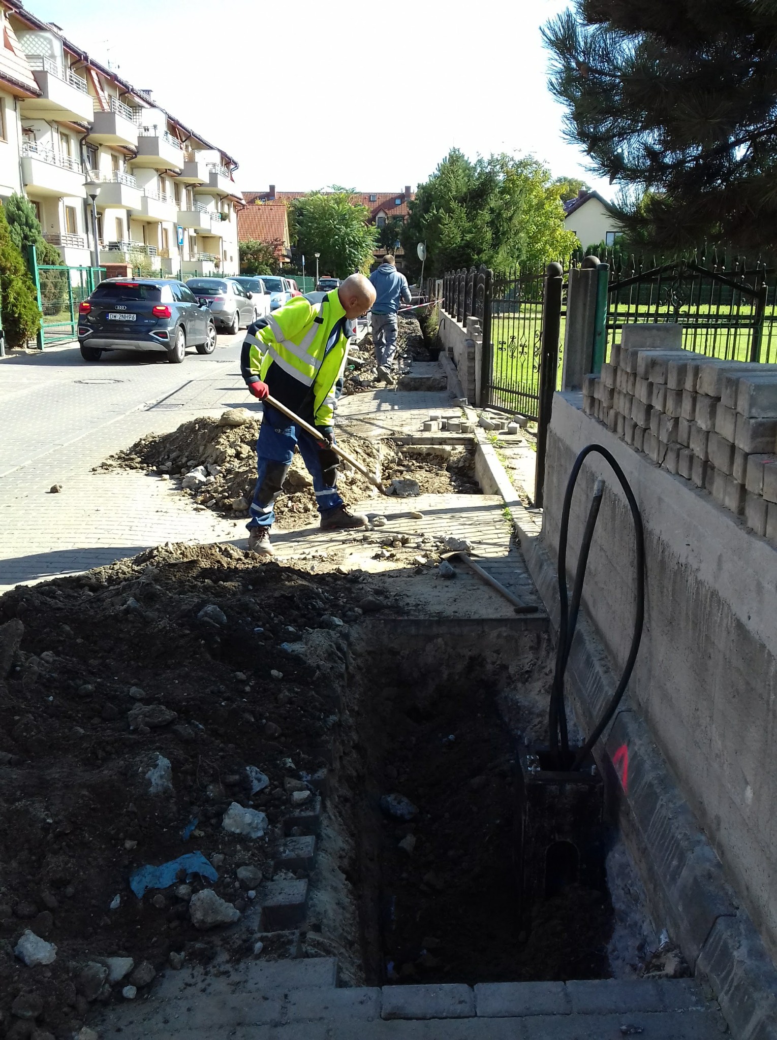 Pracownik w żółtej kamizelce kopie łopatą w wykopie obok chodnika z kostki brukowej, w tle budynki mieszkalne i zaparkowane samochody, widoczne kable w ziemi.