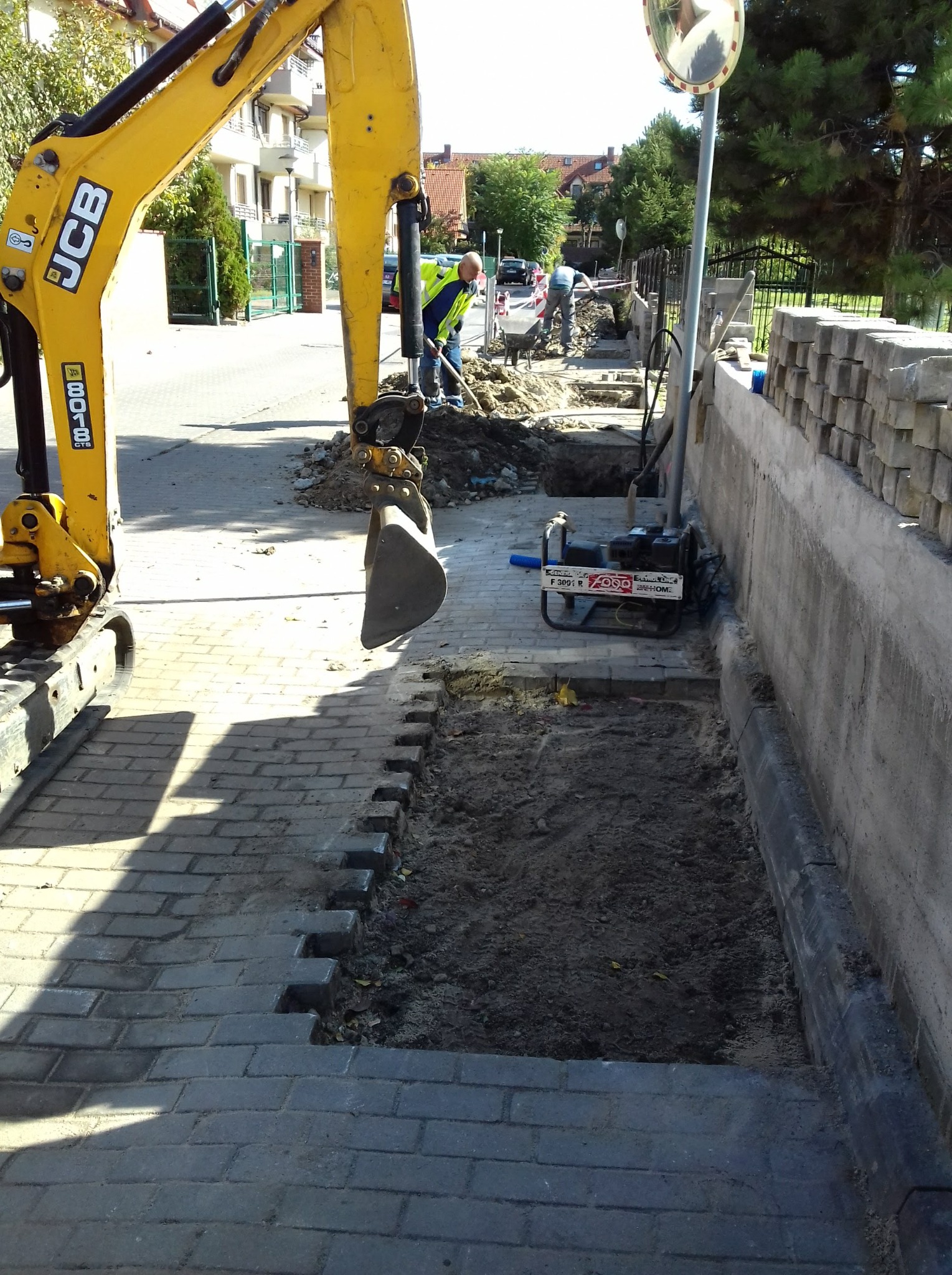 Żółta koparka JCB przy wykopie w chodniku z kostki brukowej, w tle pracownicy i sprzęt budowlany.