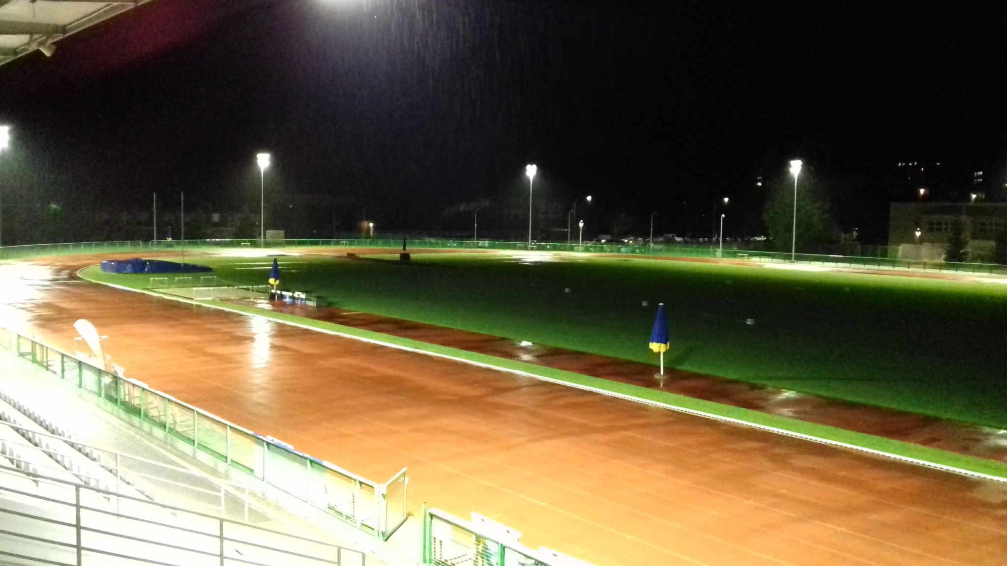 Oświetlony stadion lekkoatletyczny w deszczową noc, widok z trybun na mokrą bieżnię i zielone boisko.