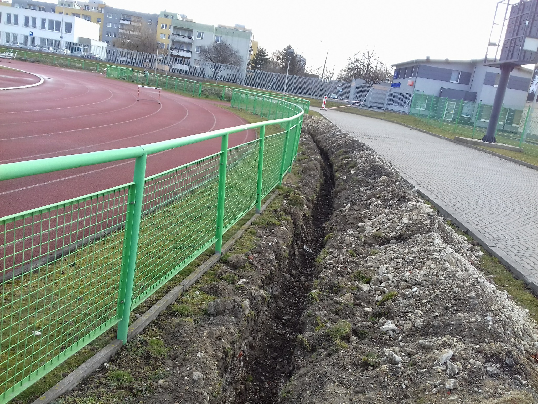 Wykopany wąski rów wzdłuż zielonego ogrodzenia boiska sportowego, obok chodnika z kostki, w tle budynki mieszkalne.