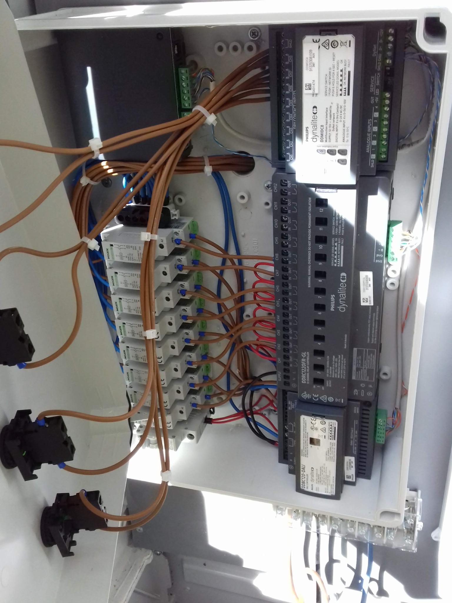 Otwarta skrzynka elektryczna z zainstalowanymi modułami Philips Dynalite, licznymi przewodami w brązowej izolacji i złączkami, widoczne elementy sterowania i zasilania.