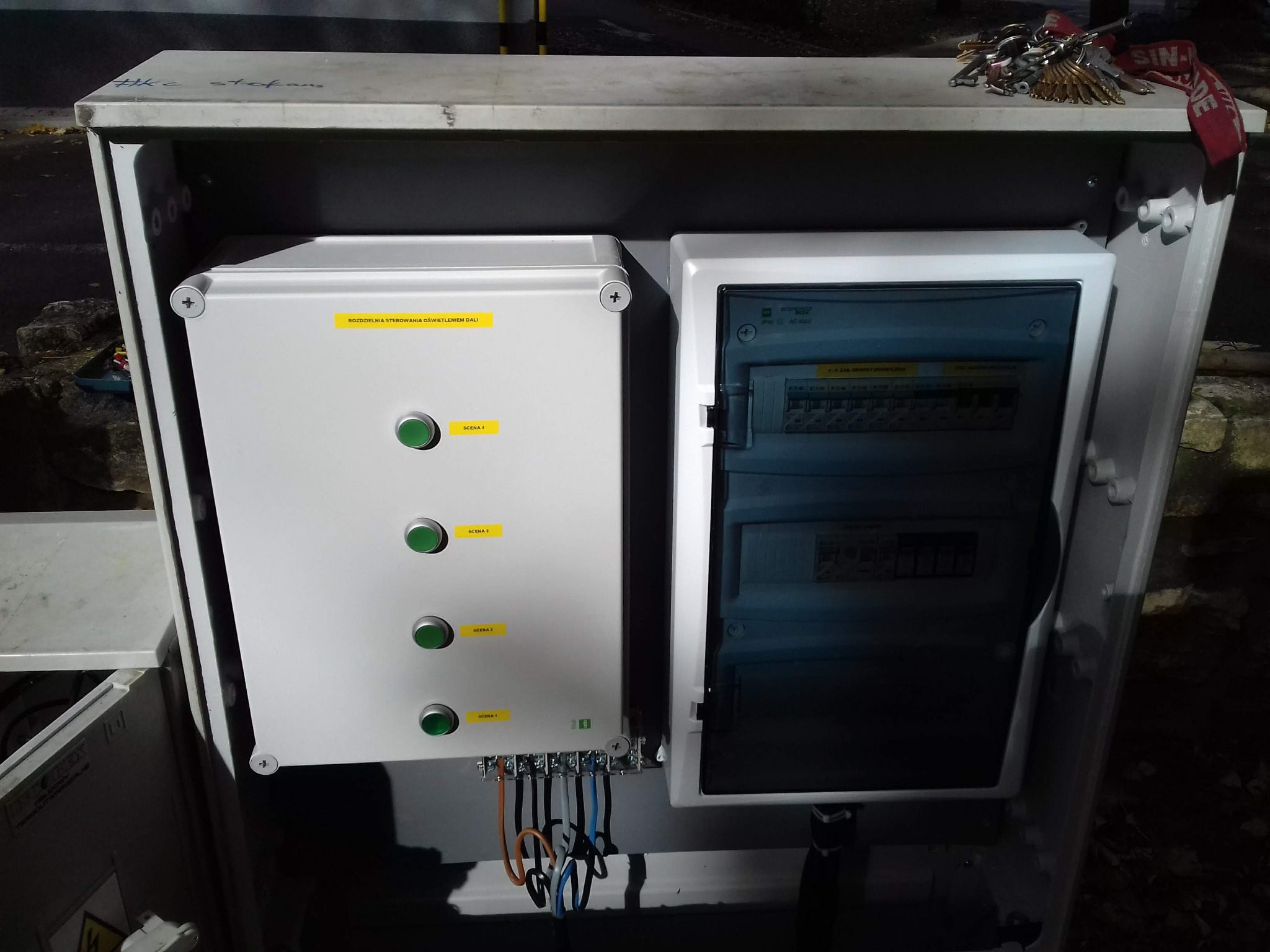 Otwarta skrzynka elektryczna z dwoma panelami: jeden z przyciskami i opisami 'Scena 1-4' oraz drugi z bezpiecznikami, klucze leżące na górze skrzynki.