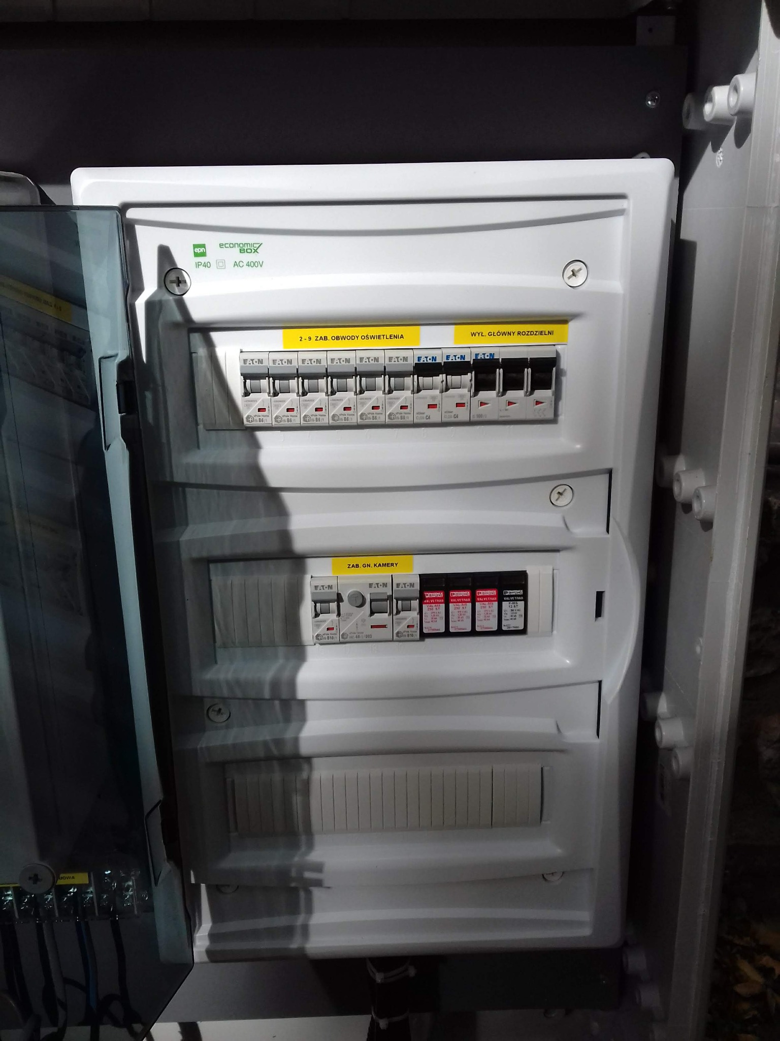 Rozdzielnia elektryczna w białej obudowie z widocznymi bezpiecznikami i oznaczeniami obwodów, umieszczona w szarej szafce, z widocznymi przewodami po lewej stronie.