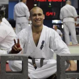 Uśmiechnięty mężczyzna w białym kimonie z czarnym pasem, siedzący na podłodze w pozycji lotosu, z dwoma betonowymi pustakami przed sobą i uniesioną ręką w geście zwycięstwa; w tle inni zawodnicy...