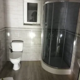 Wykończona łazienka z kabiną prysznicową z ciemnego szkła, toaletą i oknem, płytki imitujące marmur na ścianach, podłoga z paneli imitujących drewno.