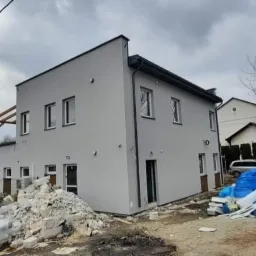 Dwukondygnacyjny budynek po tynkowaniu elewacji zewnętrznej na szaro, z widocznymi stertami gruzu i materiałów budowlanych przed budynkiem, biały samochód dostawczy w tle, pochmurne niebo.