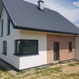 Nowoczesny dom jednorodzinny z białą elewacją, drewnianą okładziną i grafitowym dachem z blachy trapezowej, widoczne elementy wykończeniowe i fragment trawnika.