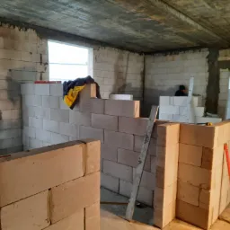 Wznoszenie ścian działowych z bloczków betonu komórkowego w surowym wnętrzu, widoczna poziomica, fragment okna i pracownik w tle.