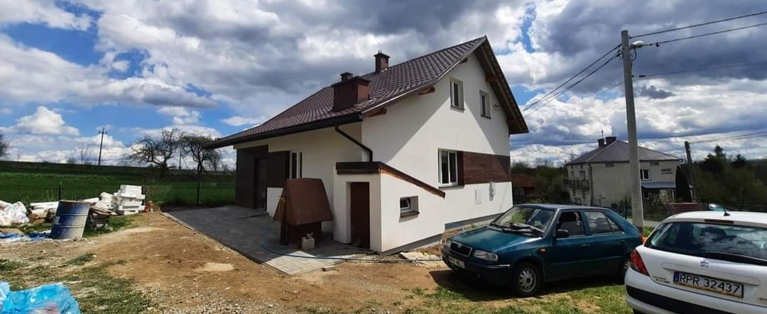 Nowoczesny dom z brązowym dachem i białą elewacją, widoczny fragment ocieplenia z brązową płytą, dwa samochody na podjeździe oraz materiały budowlane obok.