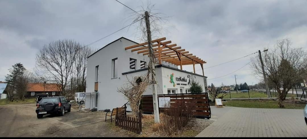 Nowoczesny budynek restauracji z jasną elewacją i drewnianą pergolą na dachu, widok z zewnątrz w pochmurny dzień. Na elewacji widoczne logo i nazwa restauracji.