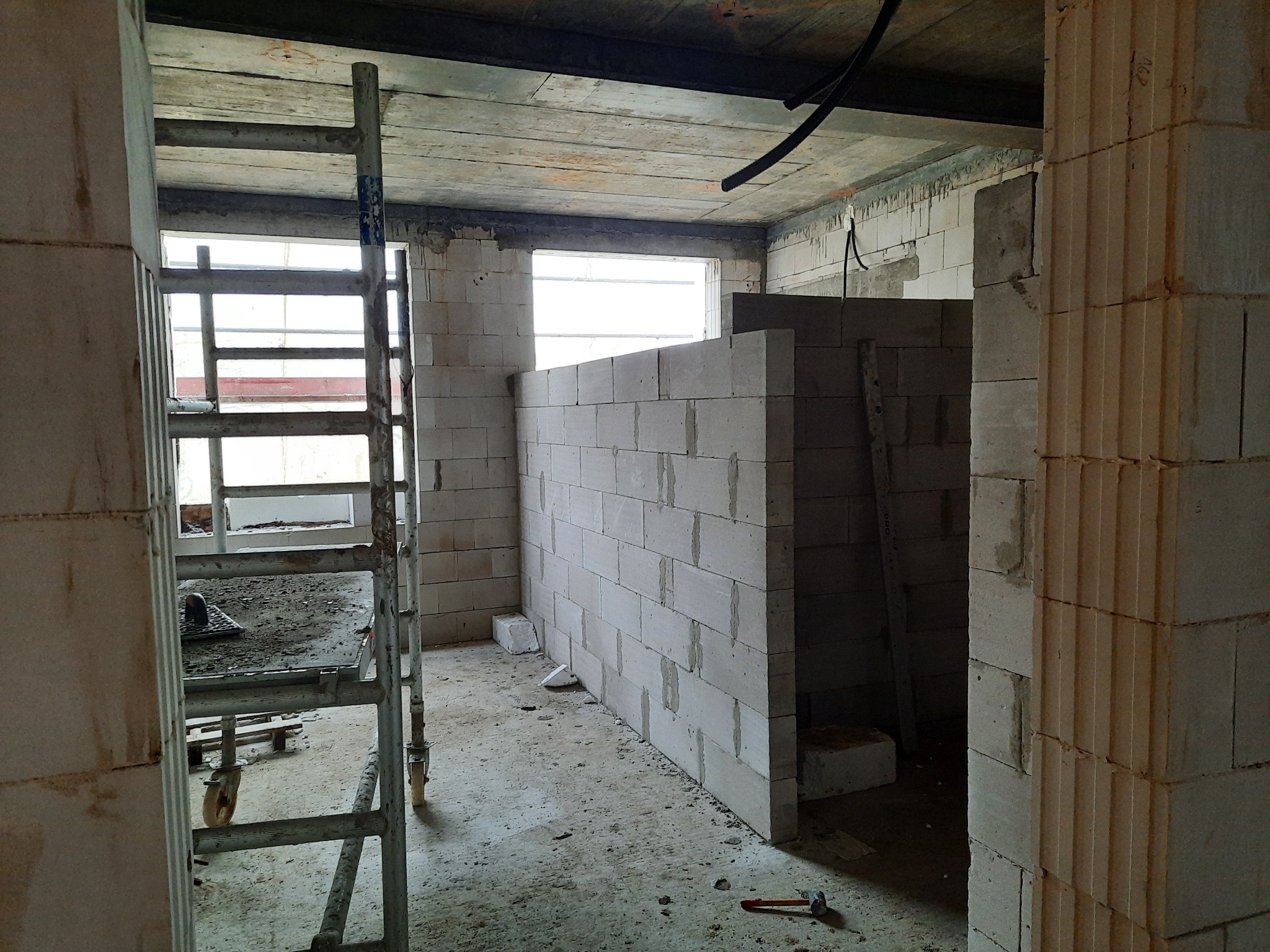 Wnętrze w trakcie budowy, ściany z bloczków betonowych, rusztowanie z narzędziami, surowy betonowy strop, instalacja elektryczna pod sufitem.