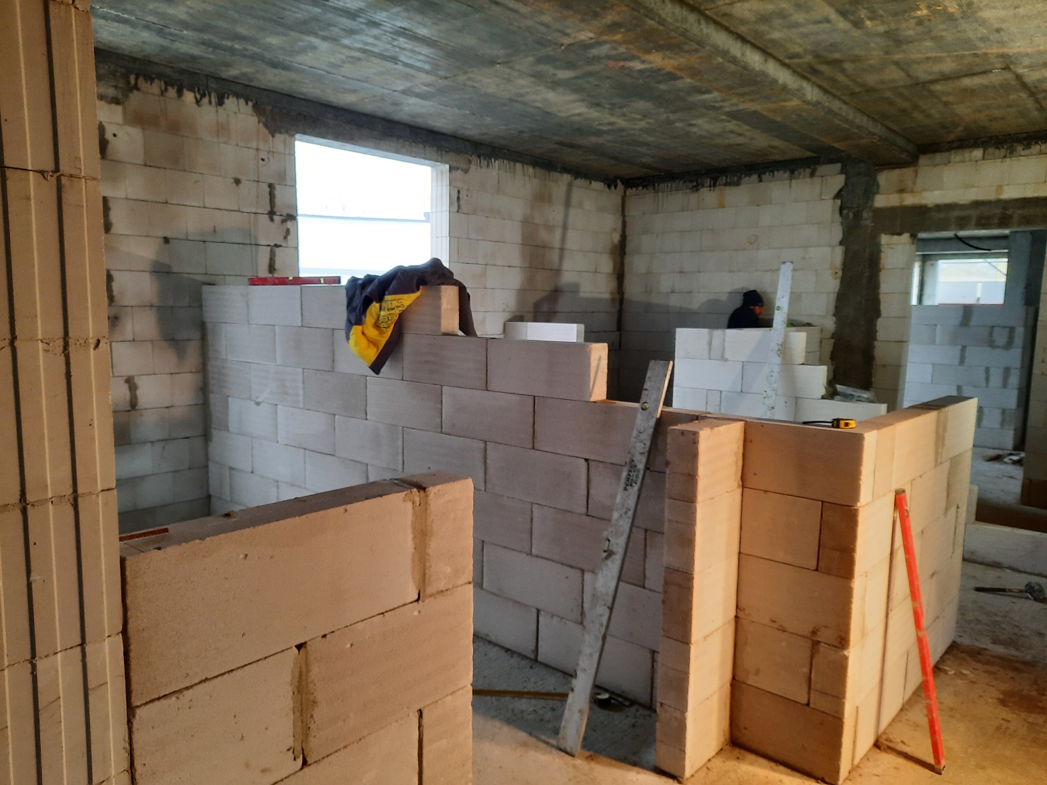 Wznoszenie ścian działowych z bloczków betonu komórkowego w surowym wnętrzu, widoczna poziomica, fragment okna i pracownik w tle.