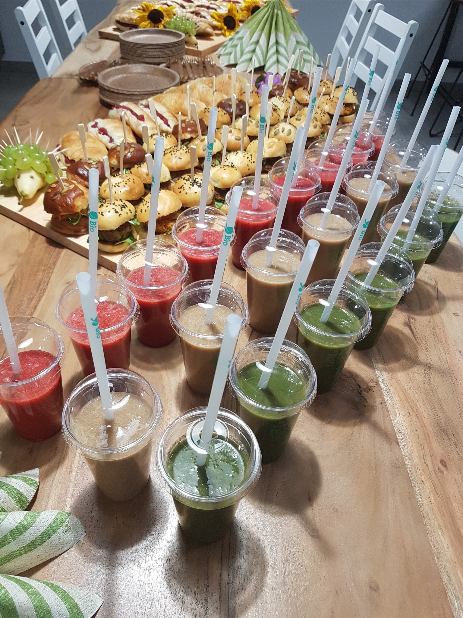 Stół z różnorodnym cateringiem: mini burgery, kanapki, przekąski i rzędy kolorowych smoothie w plastikowych kubkach z papierowymi słomkami, całość na drewnianym stole.