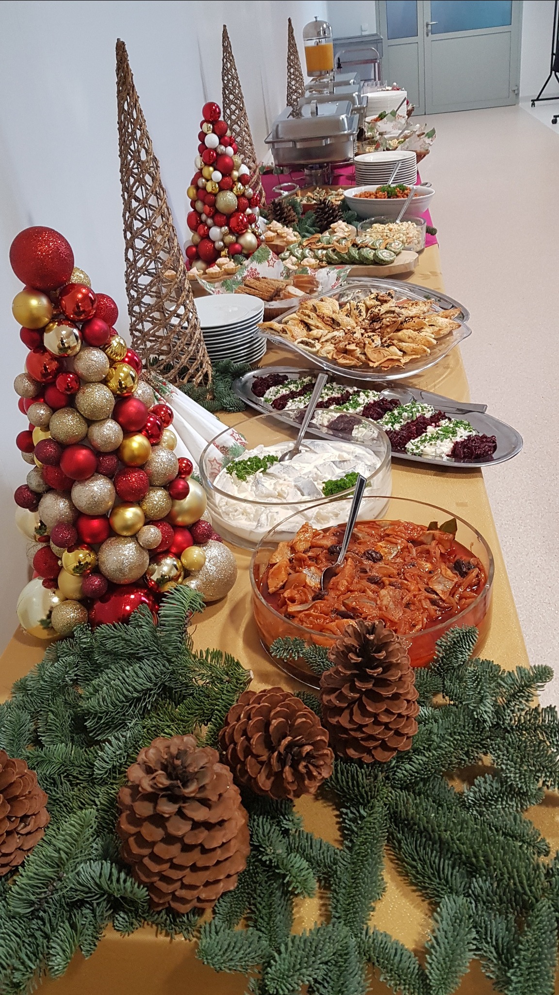 Stół cateringowy z dekoracjami świątecznymi: stroiki z bombek, gałązki świerku, szyszki oraz różnorodne potrawy w szklanych misach i na srebrnych półmiskach.