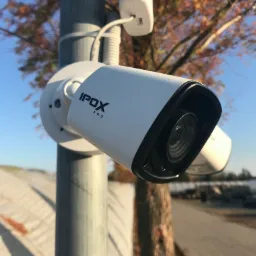 Kamera IP Ipox Pro zamontowana na metalowym słupie, z widocznym przewodem i tłem drzew i szklarni.