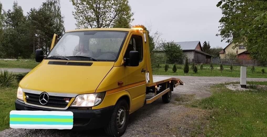 Żółta laweta Mercedes-Benz z otwartymi drzwiami kierowcy, zaparkowana na żwirowej drodze przed wiejskim domem, włączone światła awaryjne.