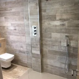 Nowoczesna łazienka z kabiną prysznicową typu walk-in, wykończona płytkami imitującymi drewno, z minimalistyczną armaturą i odpływem liniowym.