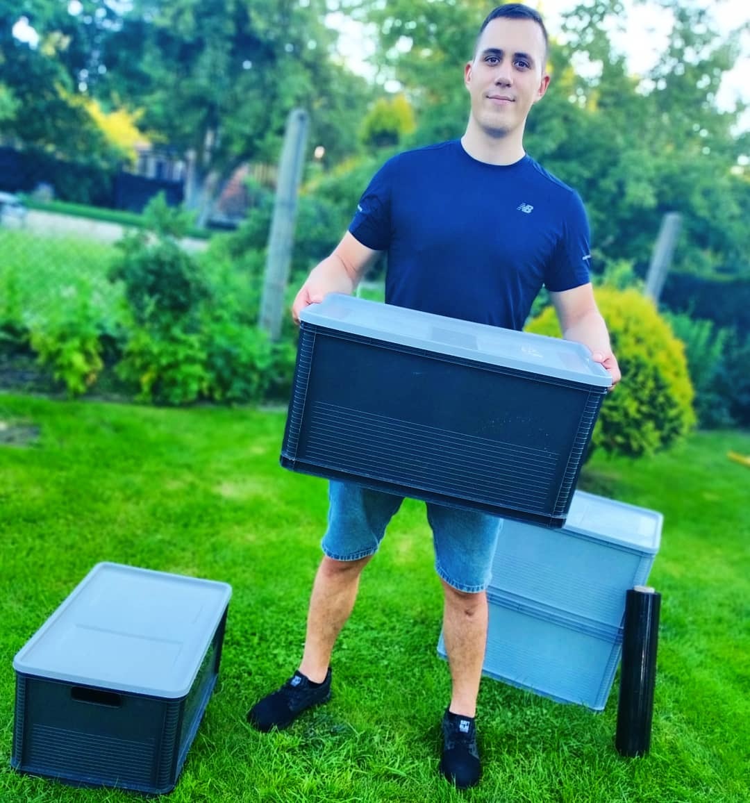 Mężczyzna w niebieskiej koszulce trzyma czarny plastikowy pojemnik z szarym wieczkiem na tle zielonego trawnika z dodatkowymi pojemnikami i folią stretch.