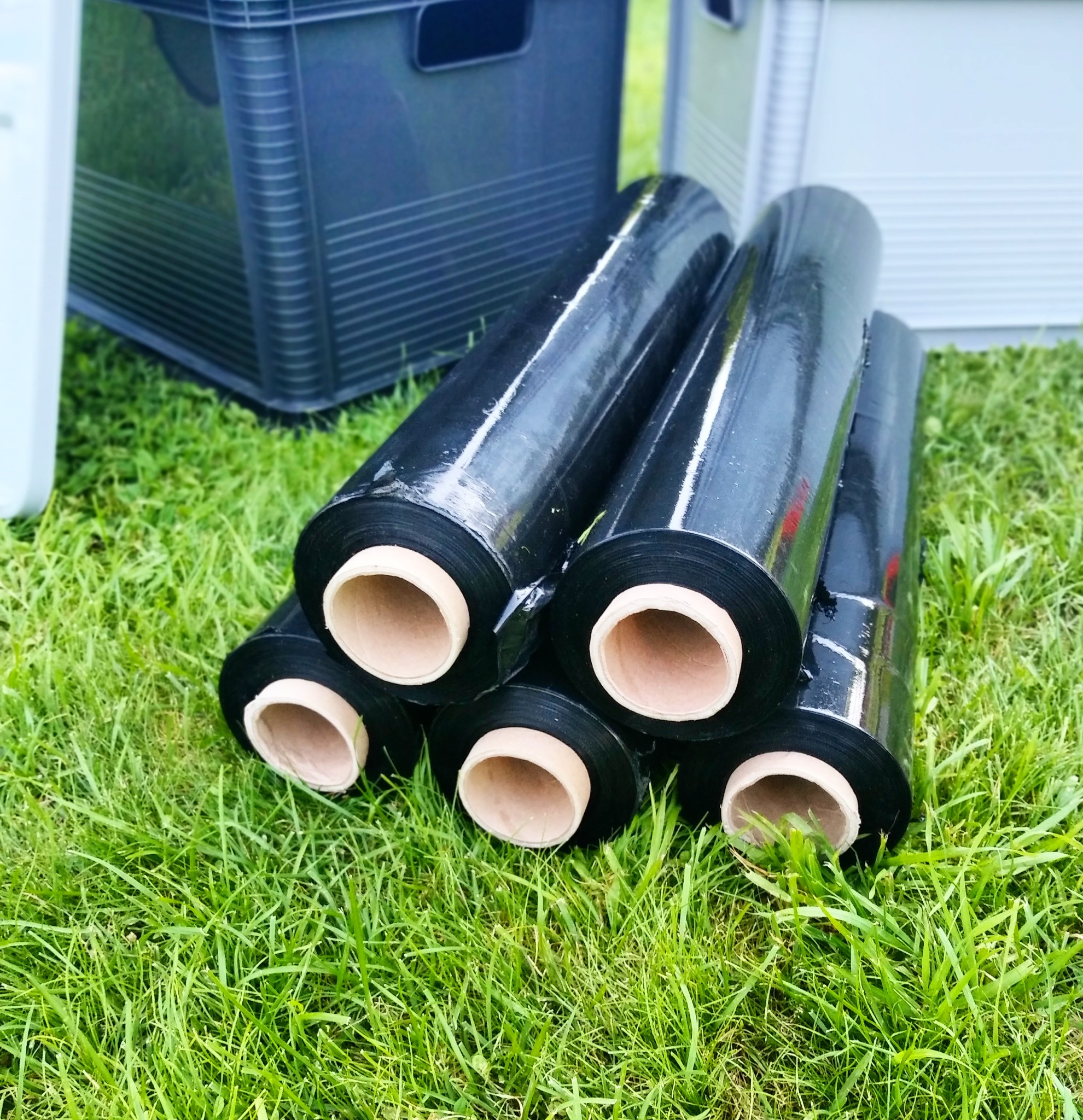 Stos czarnych rolek folii stretch z tekturowymi rdzeniami, leżący na trawie obok szarej skrzynki transportowej.