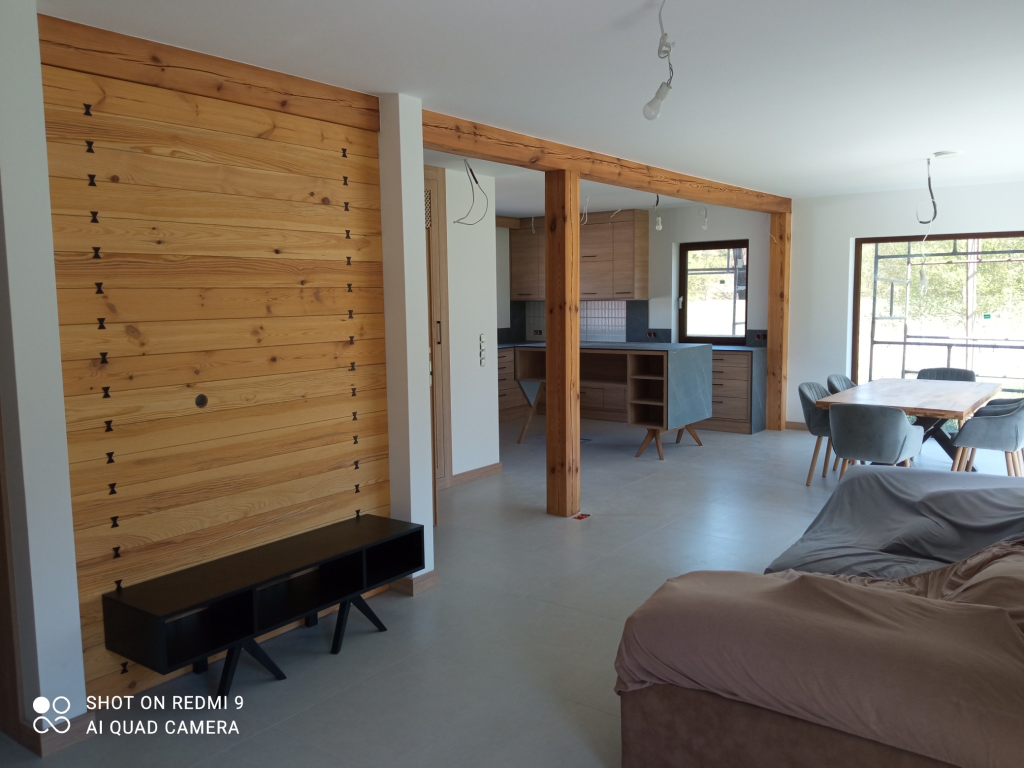 Jasne, przestronne wnętrze z drewnianą ścianą akcentującą, czarną szafką RTV i widoczną kuchnią. Drewniane belki stropowe dodają rustykalnego charakteru.