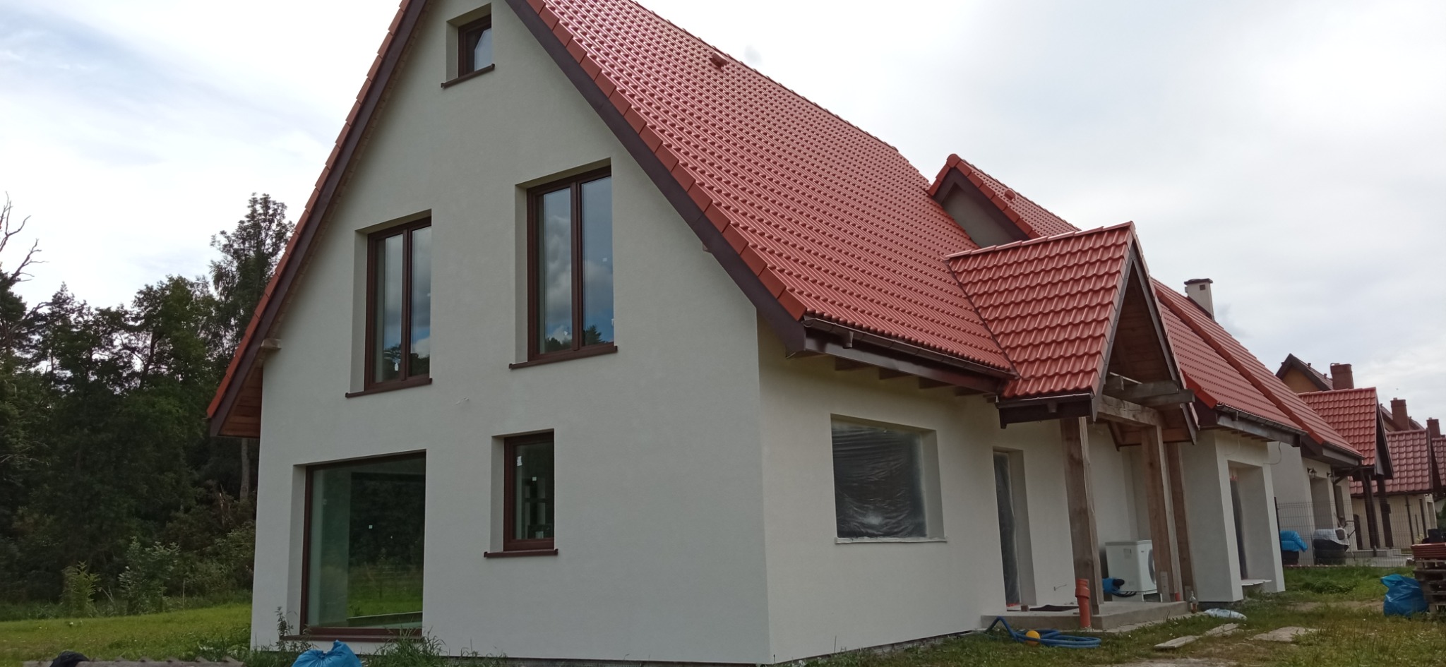 Nowo otynkowany dom jednorodzinny z czerwoną dachówką, widok z zewnątrz, z oknami z brązowymi ramami. Część okien zabezpieczona folią malarską.