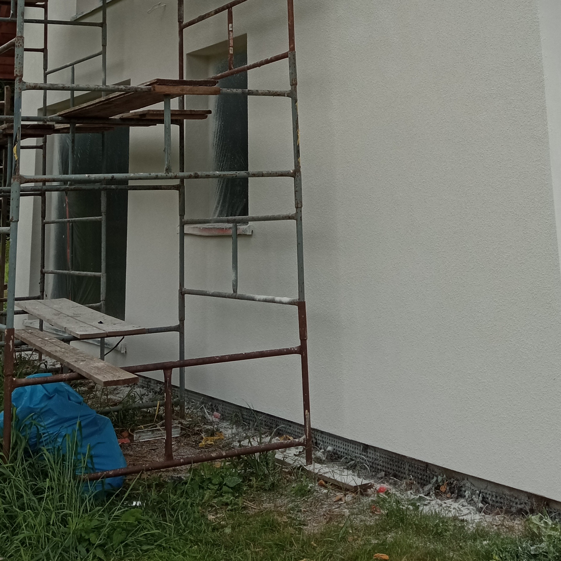 Świeżo otynkowana ściana budynku z rusztowaniem, okno zabezpieczone folią. Widoczne ślady prac remontowych na trawie.