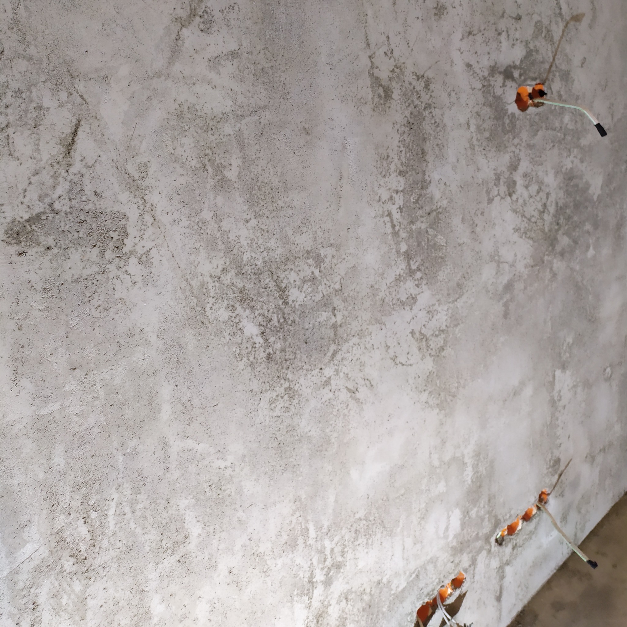 Surowa, szara ściana z widocznymi puszkami instalacji elektrycznej. W tle widoczna podłoga w trakcie prac wykończeniowych.