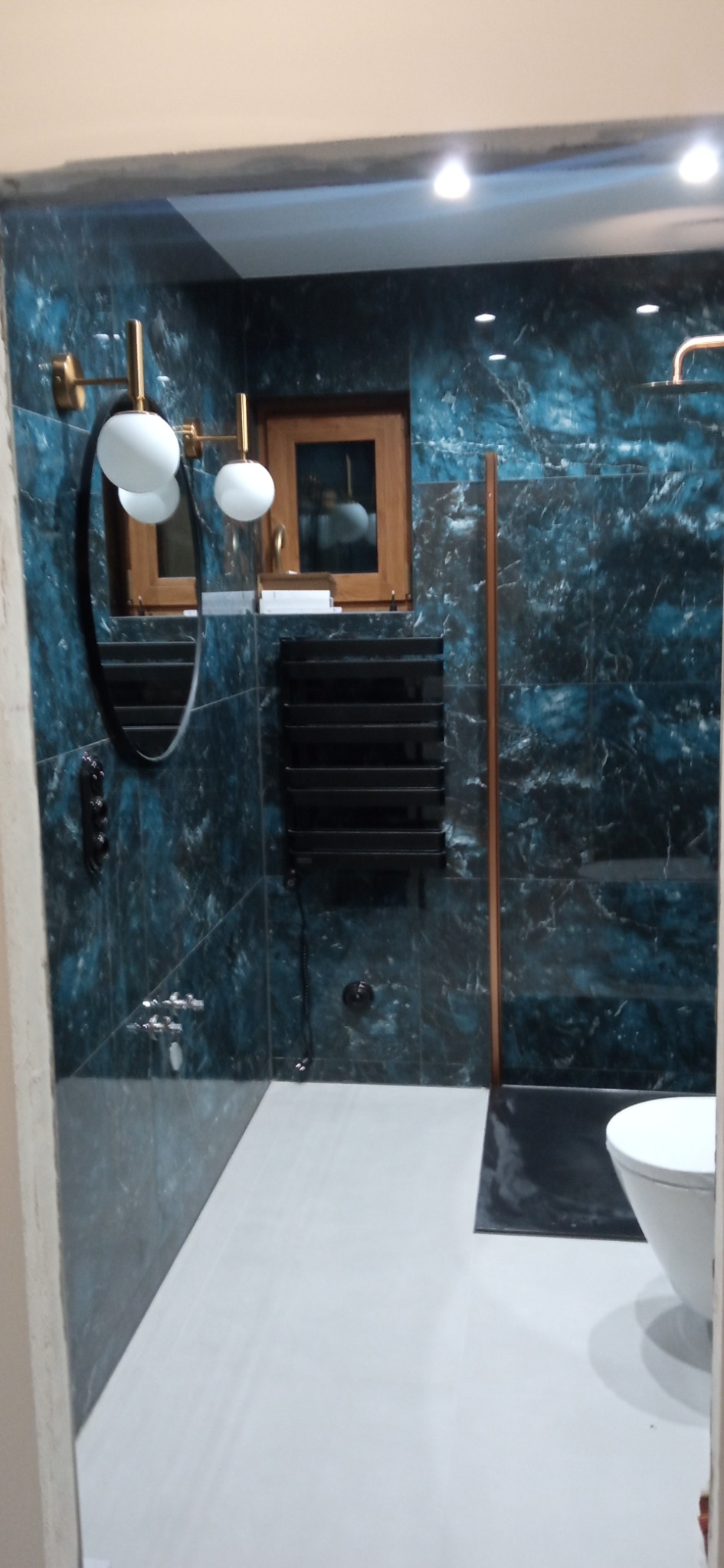 Wykończona łazienka w odcieniach granatu z marmurkowym wzorem, z owalnym lustrem i grzejnikiem drabinkowym. Widoczne białe sanitariaty i złote elementy oświetlenia.