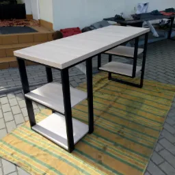 Nowoczesne biurko ze stalowym stelażem i jasnymi półkami z płyty, sfotografowane na zewnątrz na tle kostki brukowej i pasiastym dywaniku.