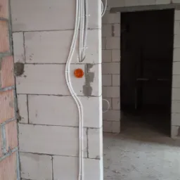 Wewnętrzna ściana z betonu komórkowego w trakcie budowy z poprowadzonymi przewodami elektrycznymi w białych peszlach i pomarańczowymi puszkami instalacyjnymi.