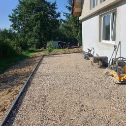 Przygotowany teren z podsypką żwirową pod budynkiem, widoczne obrzeża, ubijarka i taczka w tle, częściowo wykończony teren.