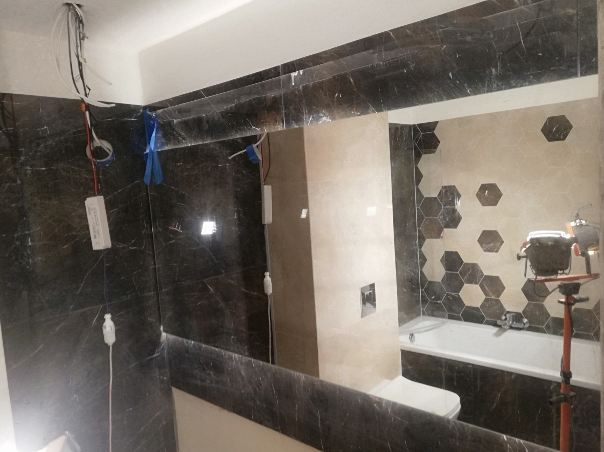 Wnętrze łazienki w trakcie prac wykończeniowych, widoczne marmurowe płytki, częściowo ułożone heksagonalne płytki na ścianie, instalacja elektryczna w suficie i lampa robocza.