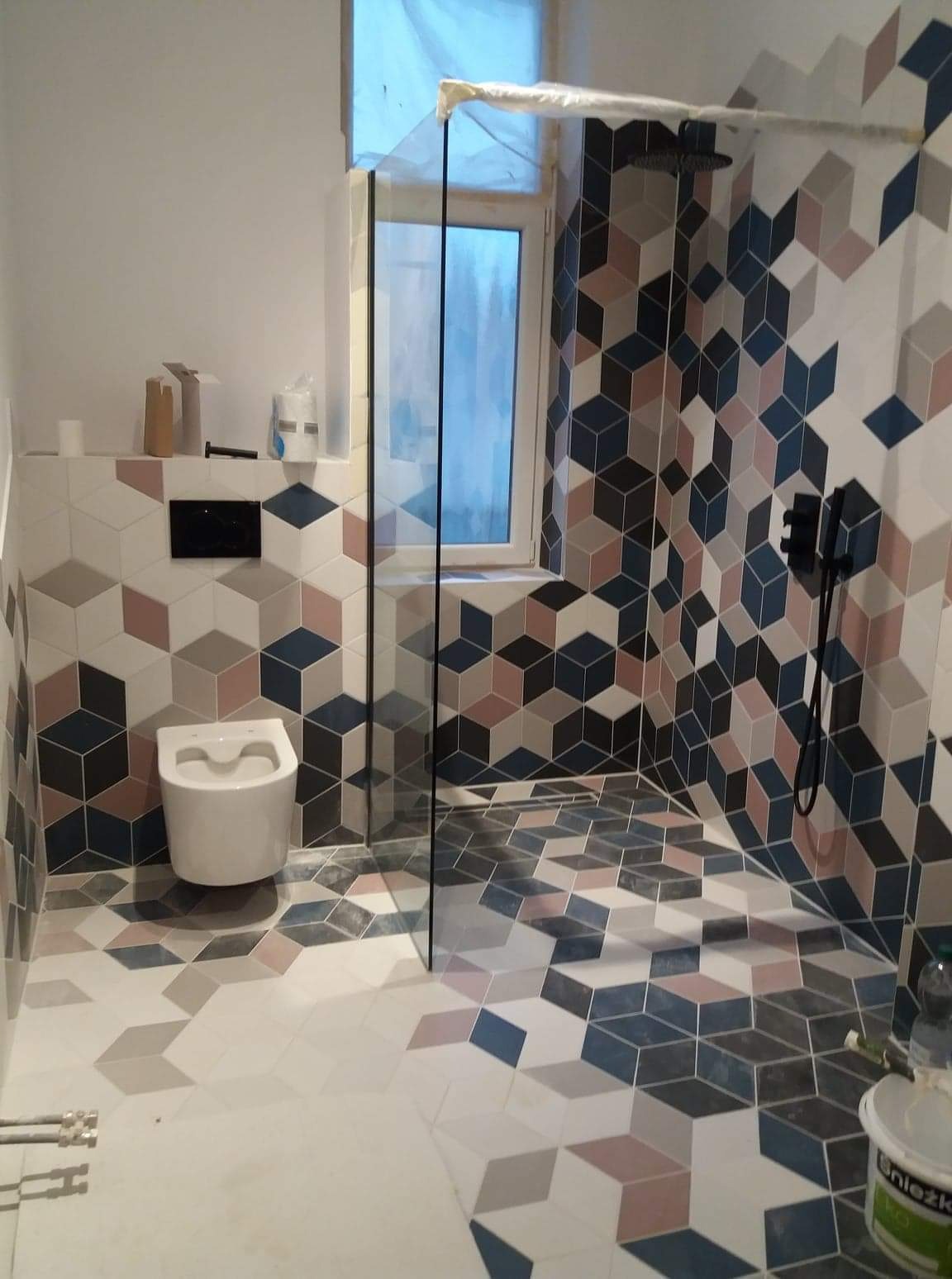 Nowoczesna łazienka po remoncie z geometrycznymi płytkami w odcieniach bieli, szarości, granatu i różu, kabina prysznicowa ze szklaną ścianką, wisząca toaleta i czarna armatura.