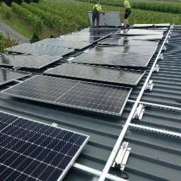 Montaż paneli słonecznych na dachu z blachy trapezowej, widoczne aluminiowe profile montażowe i pracownicy w odblaskowych ubraniach na tle zieleni.