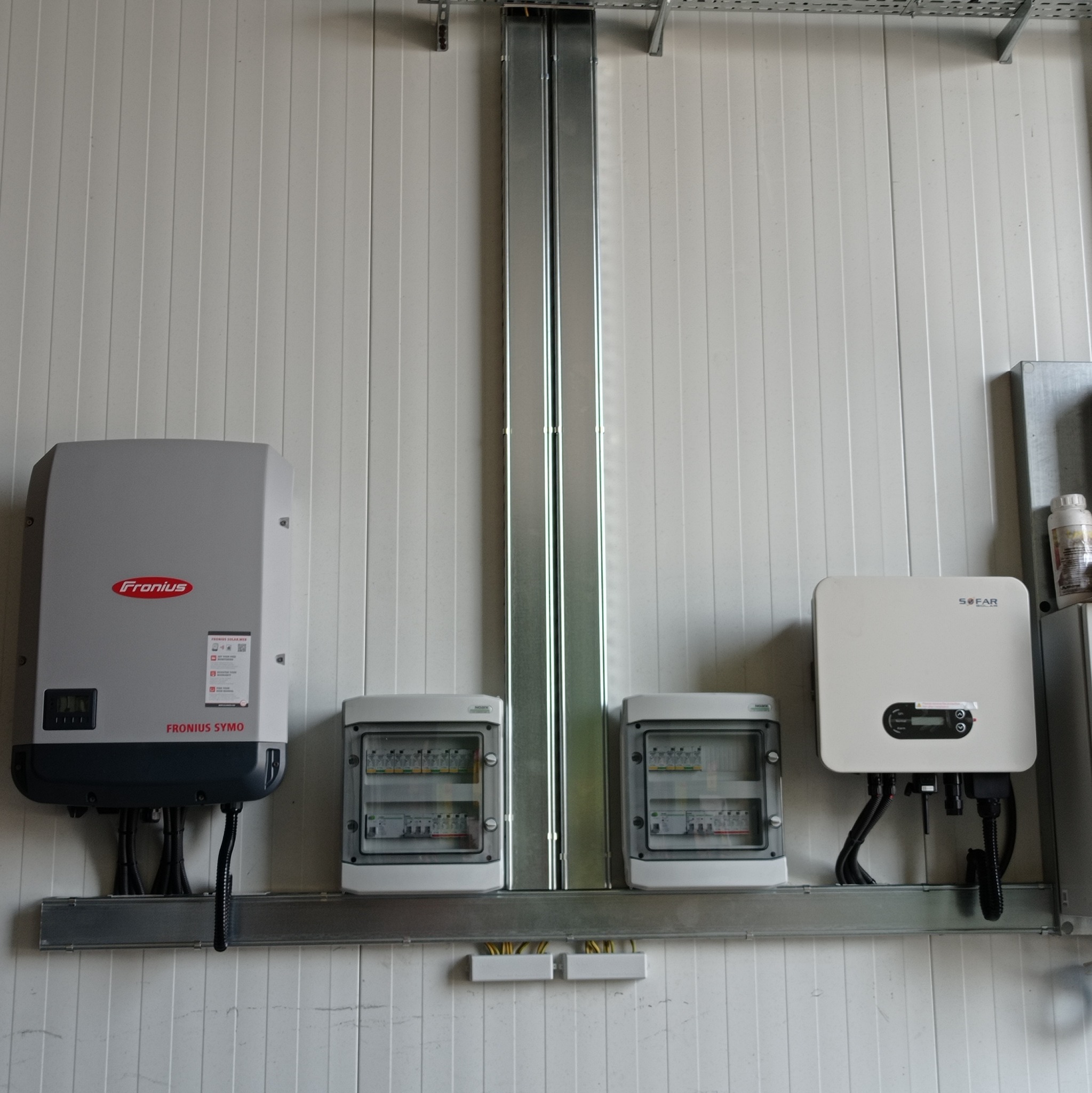 Dwa inwertery (Fronius i SOFAR) oraz dwie skrzynki elektryczne zamontowane na metalowych profilach przy ścianie z paneli. Widoczne przewody i elementy instalacji.