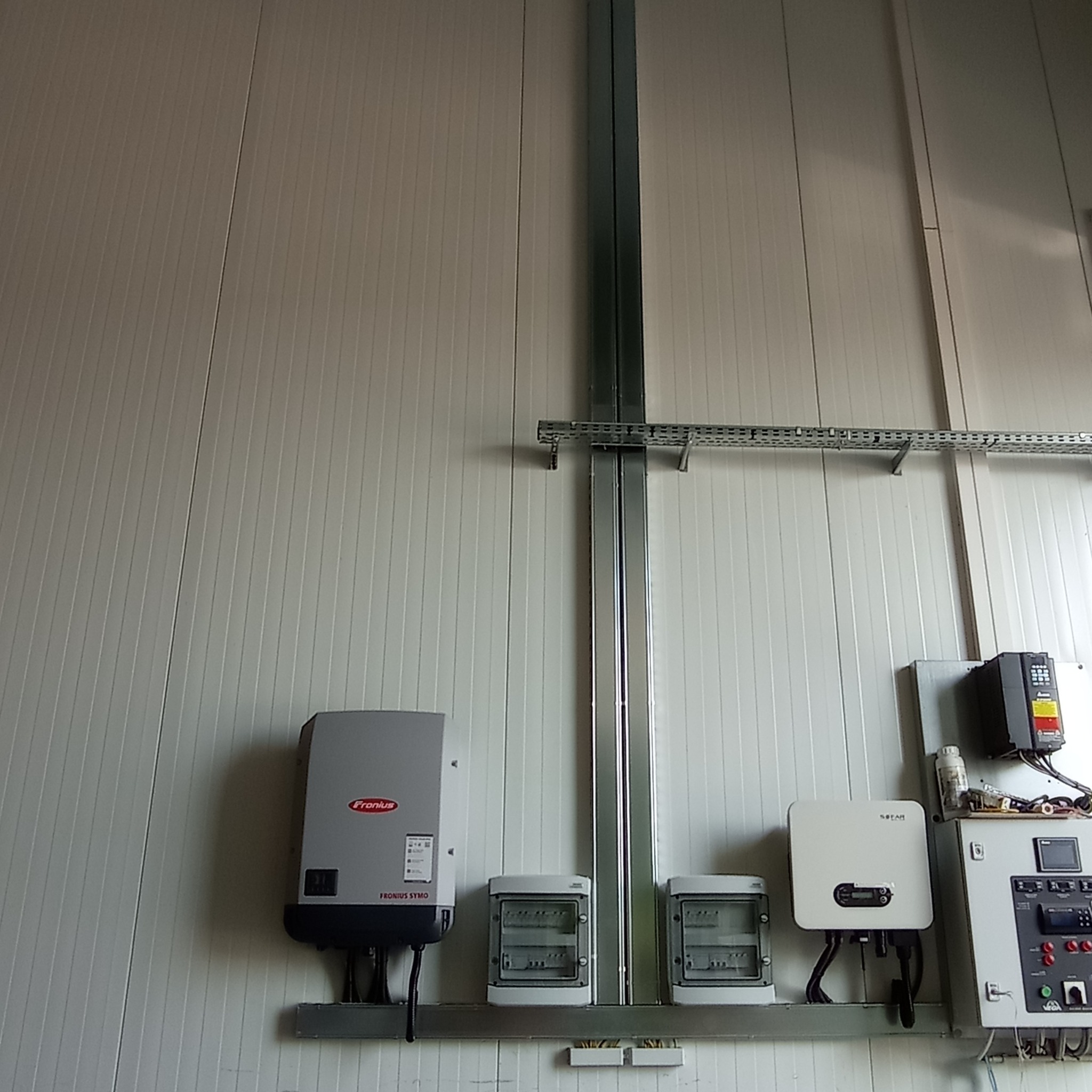 Szara ściana z panelami i metalowymi profilami podtrzymującymi skrzynki elektryczne, inwertery Fronius i Sofar oraz szafę sterowniczą.