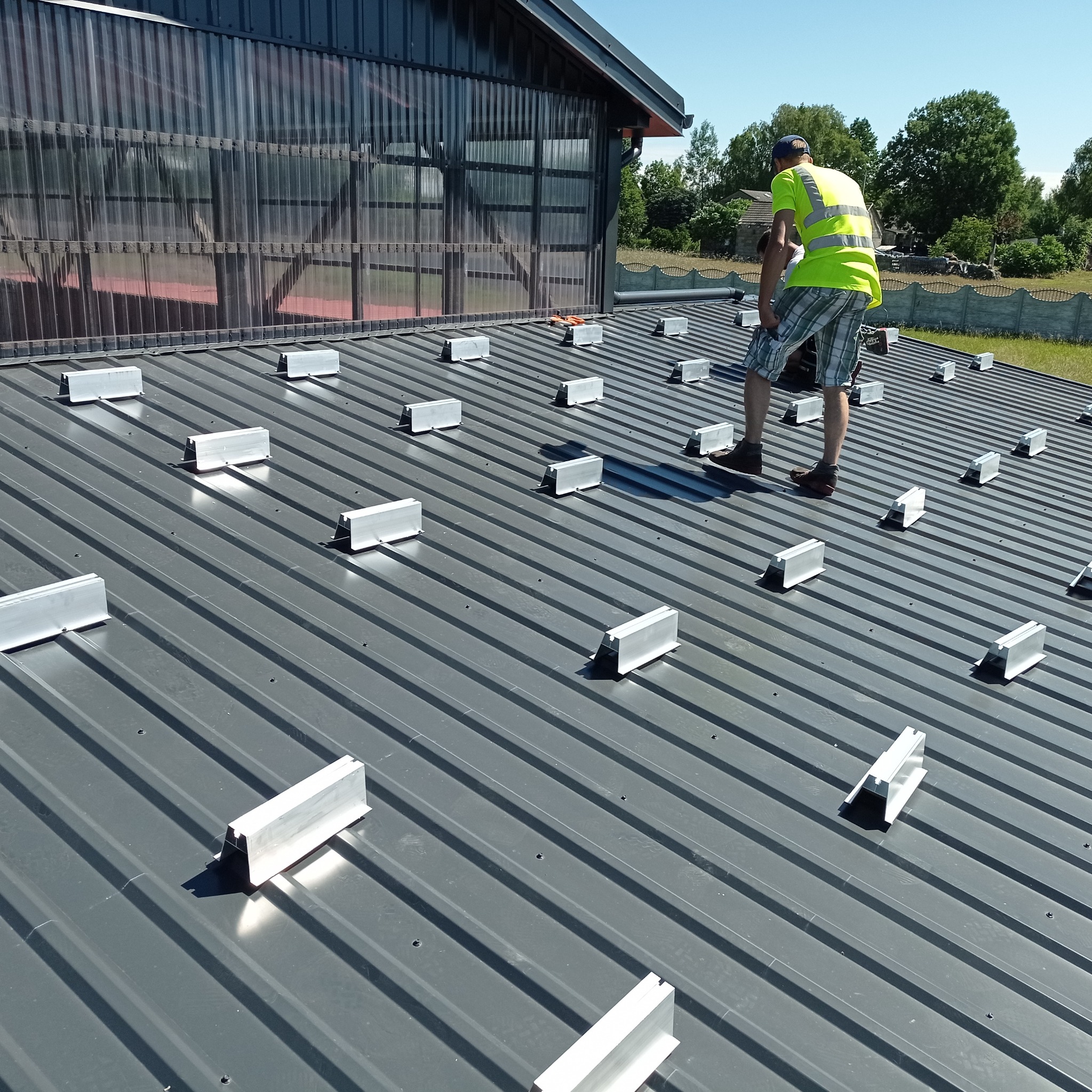 Montaż konstrukcji wsporczej pod panele fotowoltaiczne na dachu z blachy trapezowej; pracownik w odblaskowej kamizelce przykręca uchwyt, widoczne narzędzia na dachu.