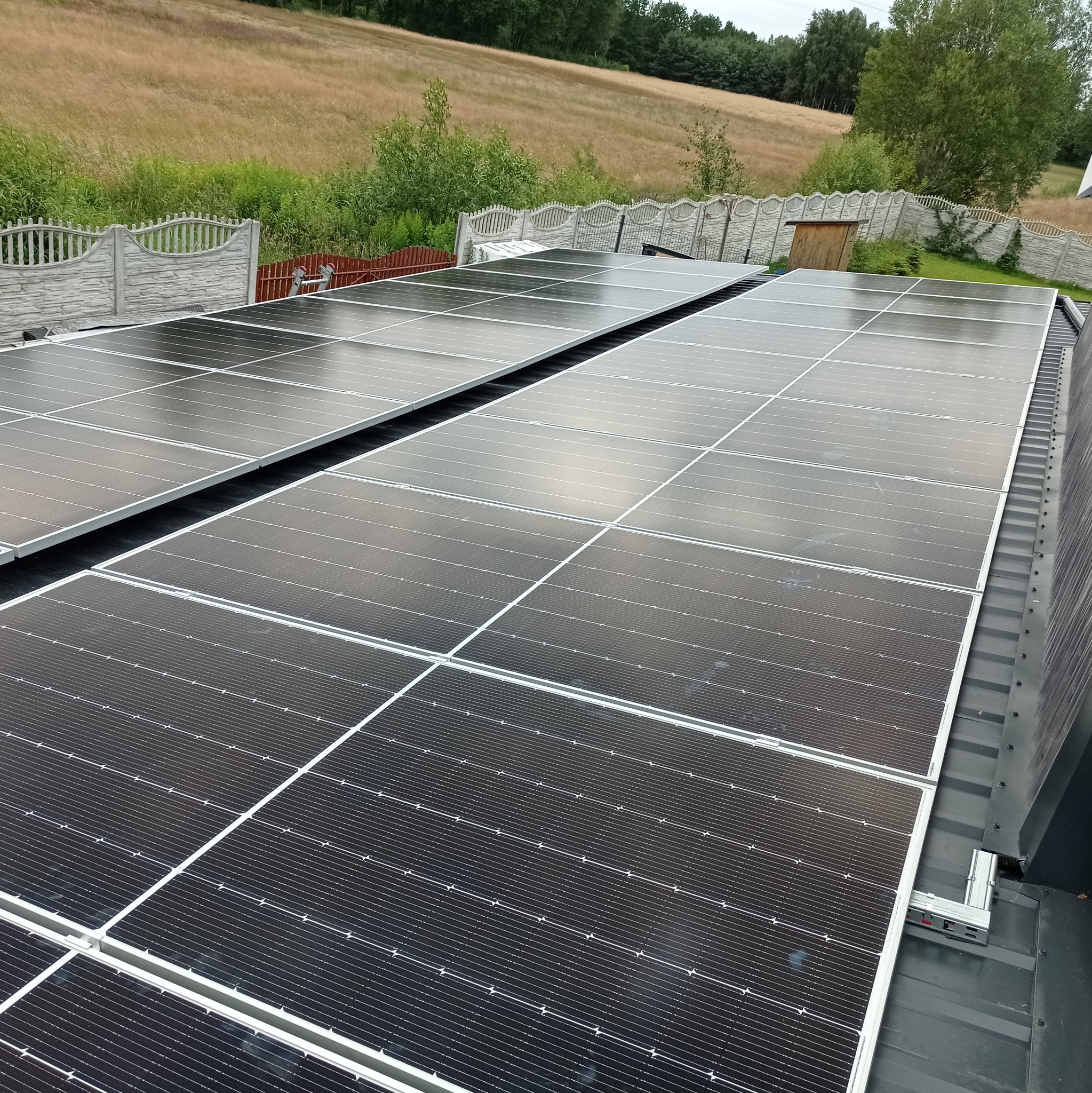 Panele słoneczne zamontowane na metalowym dachu z widokiem na wiejski krajobraz z ogrodzeniem w tle.